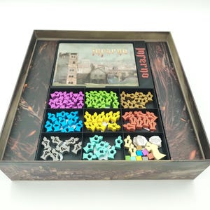 Puede incluir: Caja de juego de mesa "Inferno" con un diseño detallado en la tapa. En el interior, compartimentos contienen piezas de juego moradas, verdes, azules, amarillas y marrones. La caja está diseñada para contener los componentes del juego.