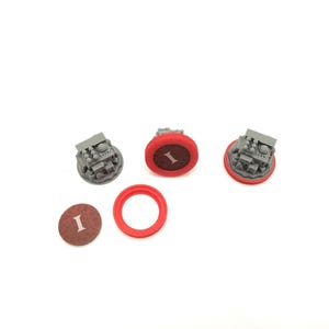 May include: Three gray miniature structures with detailed features, each surrounded by a red ring. One brown disc with the letter "I" is visible, along with a separate red ring. The items are arranged on a white surface.