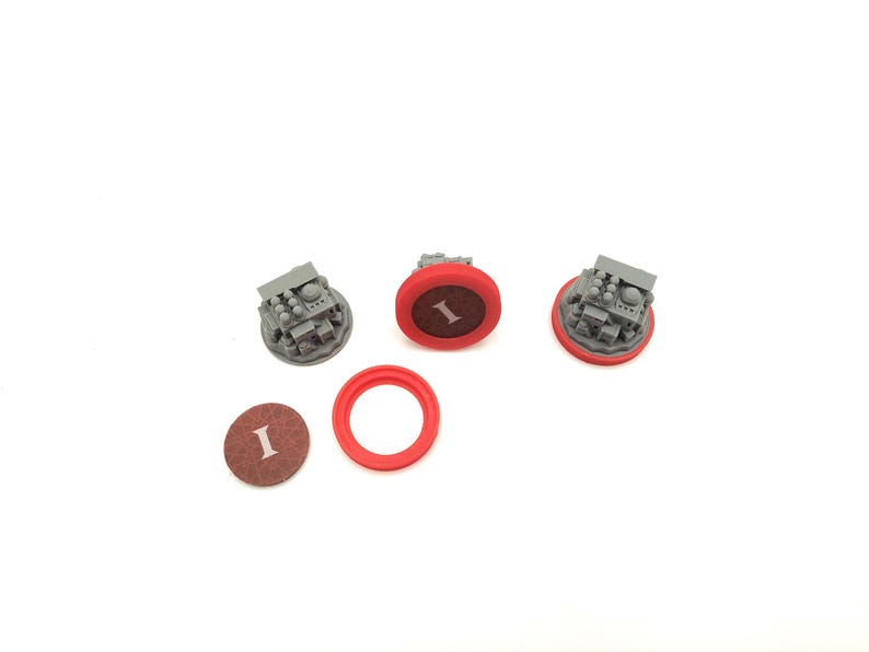May include: Three gray miniature structures with detailed features, each surrounded by a red ring. One brown disc with the letter "I" is visible, along with a separate red ring. The items are arranged on a white surface.