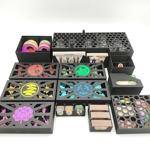 Könnte beinhalten: Eine Sammlung schwarzer, lasergeschnittener Spielorganisatoren. Die Organizer verfügen über verschiedene Fächer und Designs, darunter sechseckige Muster und dekorative Ausschnitte mit farbigen Akzenten. Das Set enthält Boxen für Karten, Token und Spielsteine.