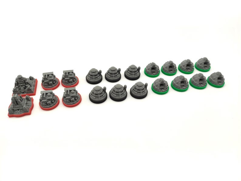 May include: A collection of miniature game pieces, including gray structures with red bases, gray dome-shaped pieces, and gray rock-like pieces with green bases. The pieces are arranged on a white surface.