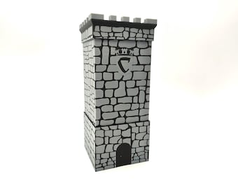 May include: A gray and black dice tower featuring a stone block pattern, a crenelated top, and a central arched doorway. The upper section displays a shield-shaped emblem with a letter C and a small crown icon above it, rendered in a high-contrast style.