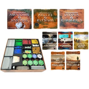 May include: A wooden game organizer with various compartments filled with game pieces in multiple colors, including green, red, blue, and yellow. Several game box covers are visible, with titles like "Amazonis & Vastitas" and "Hellas & Elysium."