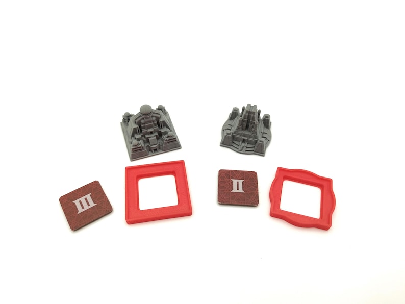 May include: A collection of board game components, including two gray miniature building models, two red square frames, and two brown square tiles with Roman numerals. The items are arranged on a white surface.