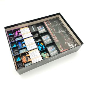 Puede incluir: Una caja negra de juego de mesa abierta, que revela compartimentos organizados. Contiene piezas de juego, cartas y un tablero. Las piezas son de varios colores, incluyendo azul, naranja y morado.