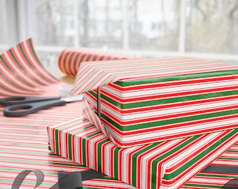 Puede incluir: Dos cajas de regalo envueltas con papel de regalo a rayas rojas, verdes y blancas. Un par de tijeras negras y un rollo de papel de regalo a juego son visibles sobre la mesa. Una cinta negra está en primer plano.