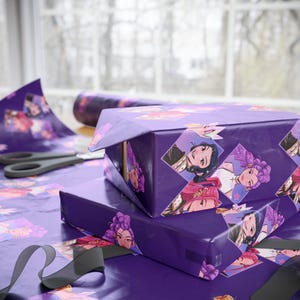 Puede incluir: Dos cajas de regalo envueltas en papel morado con ilustraciones de mujeres. El papel de regalo está decorado con retratos en forma de diamante. Se ven tijeras y un rollo de papel a juego.