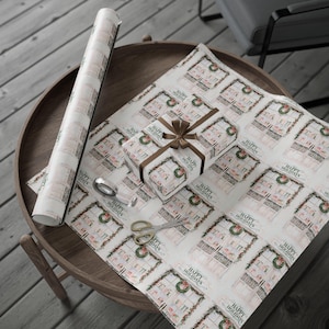 May include: A roll of wrapping paper and a wrapped gift with a brown ribbon sit on a wooden table. The wrapping paper and gift feature a repeating pattern of a window with a wreath and the words "Happy Holidays". Scissors and tape are also on the table.