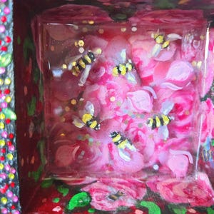 Puede incluir: Una caja decorativa con un diseño floral y un inserto cuadrado transparente. Dentro del inserto hay rosas rosas, purpurina dorada y varias abejas pequeñas con rayas amarillas y negras. La caja tiene un fondo rojo con flores pintadas.