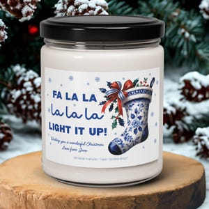May include: A glass jar candle with a black lid, displaying the text "FA LA LA LA LA LIGHT IT UP!" and a Christmas stocking illustration. The candle is placed on a wooden stand, set against a backdrop of snowy pine cones and berries.