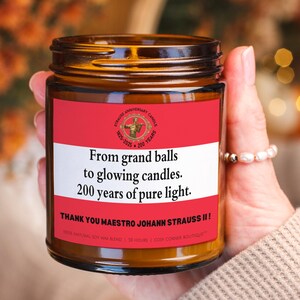 Könnte beinhalten: Eine braune Glas-Kerze mit einem rot-weißen Etikett. Auf dem Etikett steht "From grand balls to glowing candles. 200 years of pure light." und "Thank You Maestro Johann Strauss II!" Die Kerze wird in einer Hand gehalten.