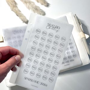 Könnte beinhalten: Eine transparente Spar-Challenge-Folie mit dem Text "50 EURO Challenge" und Kreisen mit der Aufschrift "50c". Die Folie befindet sich in einem weißen Ordner mit einem Stift. Der Text "SPARSUMME: 2000c" ist ebenfalls sichtbar.