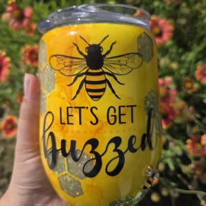 Könnte beinhalten: Gelber Becher mit einer schwarzen Bienenillustration und dem Schriftzug "LET'S GET buzzed" in schwarzer Schrift. Der Becher hat ein Wabenmuster und einen transparenten Deckel. Eine kleine Biene befindet sich am Boden.