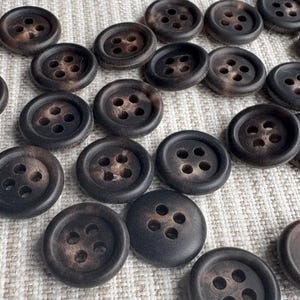May include: A collection of dark brown buttons with four holes each. The buttons have a slightly textured surface and a mottled appearance, suggesting a natural material. They are arranged on a light-colored, textured fabric.