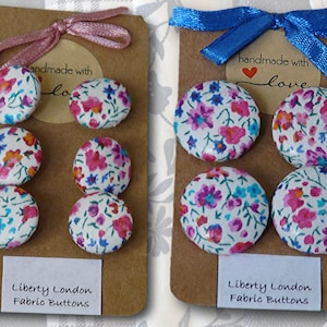 May include: Two sets of four fabric covered buttons with floral patterns in shades of pink, blue, and white. The buttons are arranged on brown card stock with a white label that reads "Liberty London Fabric Buttons" and a smaller label that reads "handmade with love".