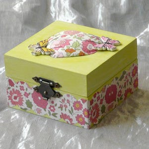 May include: A square lime green wooden box featuring a metal latch on the front. The lid is decorated with a floral pattern and two butterfly cutouts. The base of the box is wrapped in a pink, white, and green floral paper with a scalloped edge.