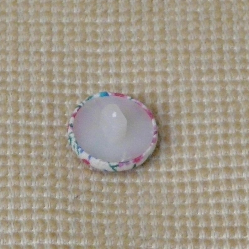 May include: A white button with a floral pattern in pink, blue, and green. The button has a clear plastic shank.