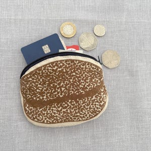 May include: A small, brown and beige patterned coin purse with a black zip, open to reveal a blue credit card and other cards. Several coins are scattered around the purse on a grey surface.