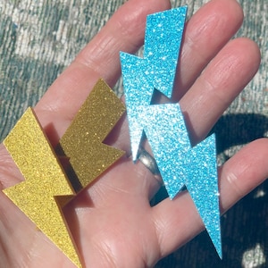 May include: Two glitter lightning bolt shapes, one gold and one blue, are held in a hand.