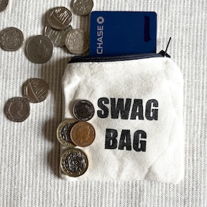 May include: A small, off-white canvas "SWAG BAG" with a black zipper and the words "SWAG BAG" printed in black. A blue credit card is partially visible inside the bag. Several coins are scattered around the bag.