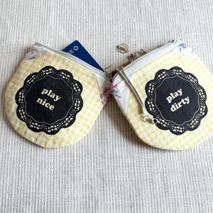 May include: Two small, yellow gingham coin purses with white zippers. One purse has a blue card peeking out. Each purse has a black, scalloped label with the phrases "play nice" and "play dirty" in white text.