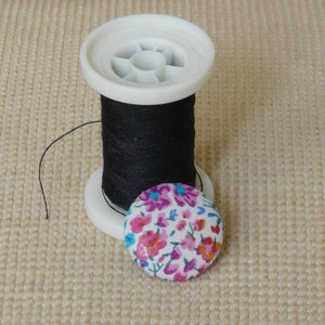May include: A spool of black thread on a white plastic bobbin with a floral fabric button.