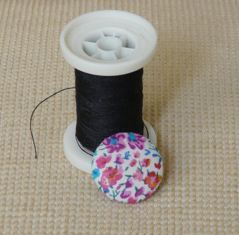 May include: A spool of black thread on a white plastic bobbin with a floral fabric button.