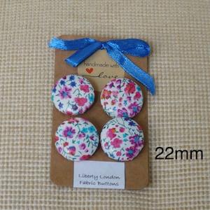 May include: Four 22mm fabric covered buttons with a floral print in shades of pink, blue, and white. The buttons are arranged in a square on a brown card with a blue ribbon and a tag that says "handmade with love". The card also says "Liberty London Fabric Buttons".