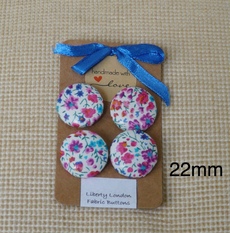 May include: Four 22mm fabric covered buttons with a floral print in shades of pink, blue, and white. The buttons are arranged in a square on a brown card with a blue ribbon and a tag that says "handmade with love". The card also says "Liberty London Fabric Buttons".