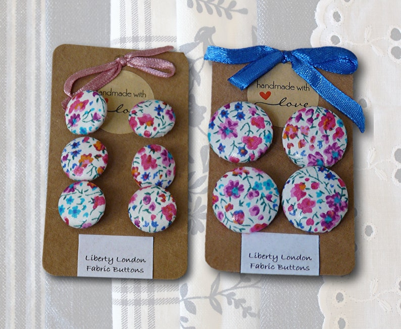 May include: Two sets of four fabric covered buttons with floral patterns in shades of pink, blue, and white. The buttons are arranged on brown card stock with a white label that reads "Liberty London Fabric Buttons" and a smaller label that reads "handmade with love".