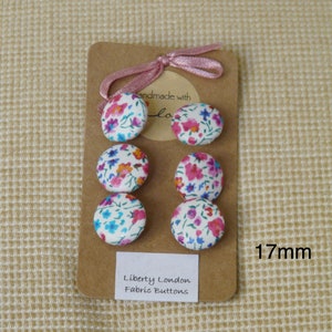 May include: Six 1.7 inch round fabric buttons with a white background and a floral pattern in pink, blue, and green. The buttons are arranged in two rows of three on a brown card with the text "Handmade with Love" and "Liberty London Fabric Buttons".