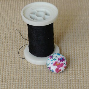 May include: A spool of black thread on a white plastic spool with a floral button.