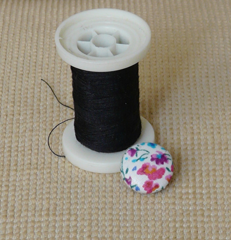 May include: A spool of black thread on a white plastic spool with a floral button.