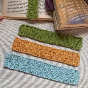 May include: Four knitted bookmarks in different colours and patterns. The bookmarks are green, orange, light blue, and purple. The bookmarks are lying on top of a book.