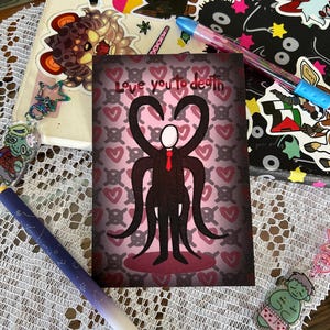 May include: A greeting card with the text "Love you to death" features a stylized figure with heart-shaped arms and a red tie, set against a background of hearts. The card is surrounded by pens, stickers, and a charm on a lace surface.