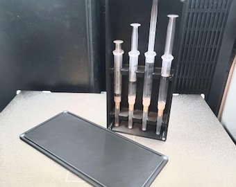 May include: A black plastic case with four syringes inside, and a separate black rectangular lid. The syringes are clear with orange tips and are held upright in the case. The lid is lying flat next to the case.