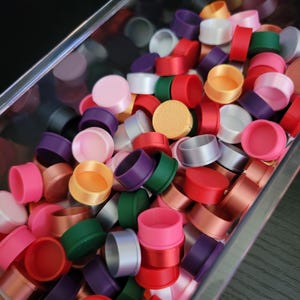 May include: A clear container filled with colorful ribbon spools. The spools are in various colors, including red, pink, purple, green, and gold. The spools are made of satin ribbon and are stacked together. The container is sitting on a dark surface.