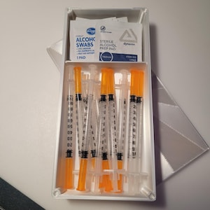 May include: A white plastic case containing several syringes with orange caps and clear barrels. The case also holds alcohol swabs and prep pads. The syringes are marked with measurements. The case is open, revealing the contents.