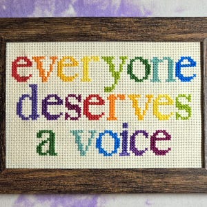 May include: Cross-stitch artwork in a brown wooden frame. The text "everyone deserves a voice" is stitched in rainbow colors on a cream-colored fabric. The background is a soft purple and white pattern.