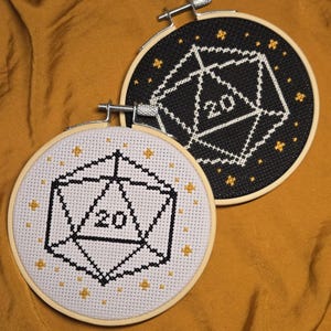 Pode incluir: Duas peças de ponto cruz em aros de madeira. Uma tem fundo preto com um dado branco e estrelas douradas. A outra tem fundo branco com um dado preto e estrelas douradas. Ambos os dados mostram o número 20.