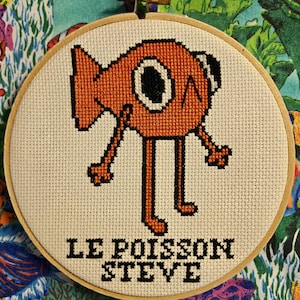 Puede incluir: Un bastidor de bordado con un pez naranja de dibujos animados, detalles negros y el texto "LE POISSON STEVE" debajo. El bastidor se muestra sobre un fondo de tela estampada vibrante. El pez tiene un ojo grande.
