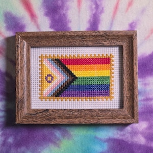 May include: A rectangular cross-stitch embroidery featuring the Progress Pride flag, framed in a dark brown wood frame. The design includes horizontal stripes of red, orange, yellow, green, blue, and violet, with a chevron pattern of white, pink, light blue, brown, and black, and a small purple circle on the left side.