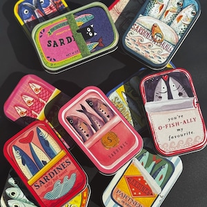 Puede incluir: Una colección de latas de sardinas rectangulares de metal con ilustraciones coloridas de peces, gatos y textos que incluyen "Sardines salty but sweet", "you're O-FISH-ALLY my favourite" y "Sardines Partners in brine" dispuestas de forma dispersa.