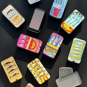 May include: A collection of rectangular metal tins featuring various colorful illustrations of sardines and anchovies, including some with text like SARDINES DE LA MER, SARDINES POUR LES TOASTS, and ANCHOVY, alongside one tin with a black and white honeycomb pattern.