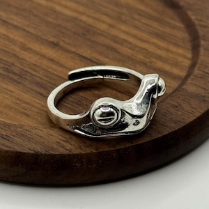 Op de afbeelding: Een zilverkleurige verstelbare ring met een mechanisch ontwerp. De ring heeft een gedetailleerde, industrieel geïnspireerde esthetiek met klinknagels en een gebogen, open band. De ring wordt gepresenteerd op een houten oppervlak.