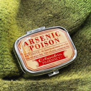 May include: A small, rectangular, silver pillbox with a hinged lid. The label inside reads "ARSENIC POISON" in red text, with additional text about an antidote. The box is set against a green, textured background.