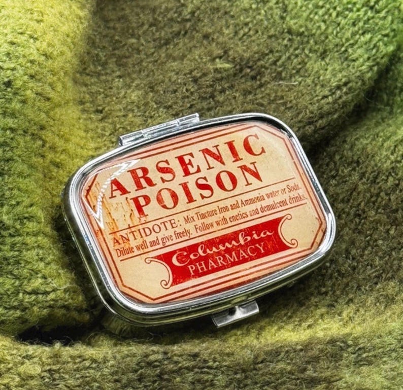 May include: A small, rectangular, silver pillbox with a hinged lid. The label inside reads "ARSENIC POISON" in red text, with additional text about an antidote. The box is set against a green, textured background.