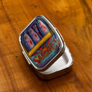 May include: A silver pill box with a hinged lid. The lid displays a colourful illustration of sardines in a can, with the word "Sardines" and "Pure Olive Oil" printed on the lid. The box is open, revealing a white interior.