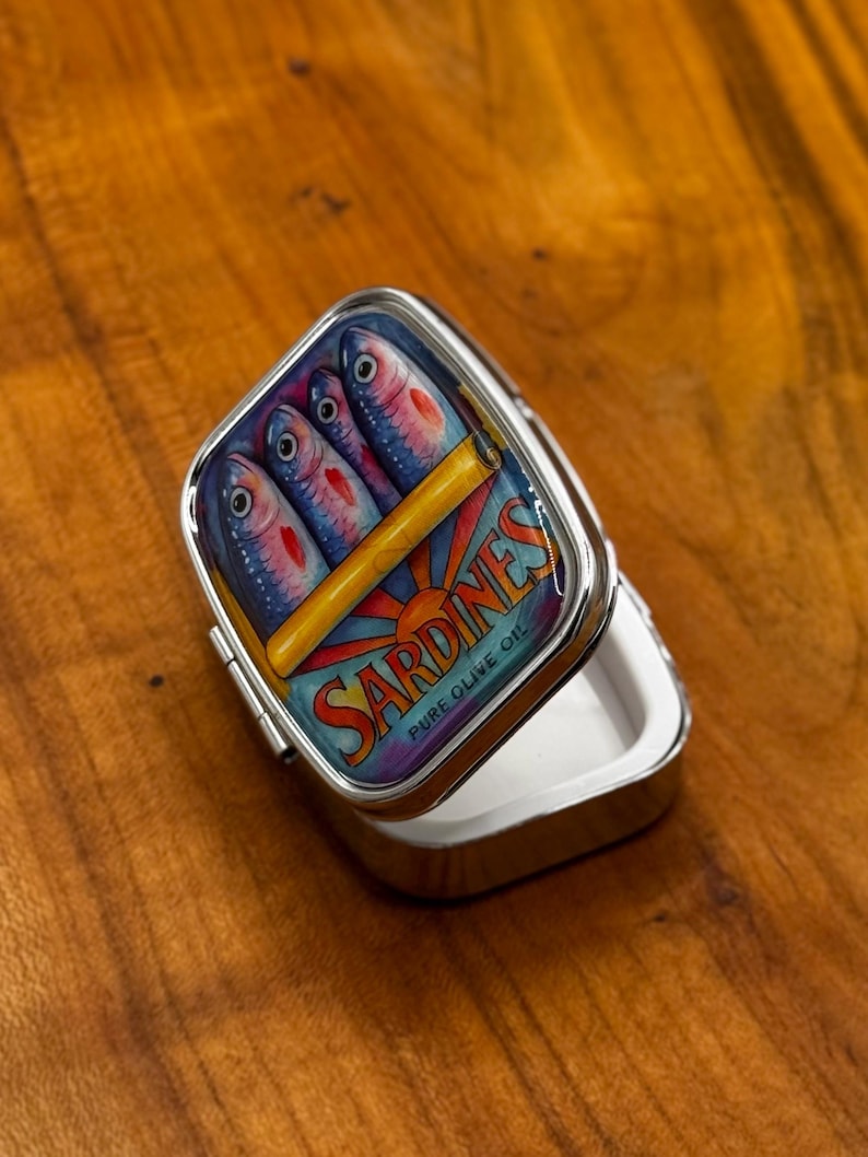 May include: A silver pill box with a hinged lid. The lid displays a colourful illustration of sardines in a can, with the word "Sardines" and "Pure Olive Oil" printed on the lid. The box is open, revealing a white interior.