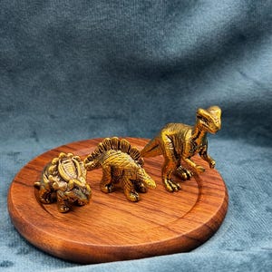 May include: Three small, metallic gold-colored dinosaur figurines standing on a circular wooden base. The figures include a triceratops, a stegosaurus, and a bipedal theropod, each featuring detailed textured skin and distinct anatomical features.